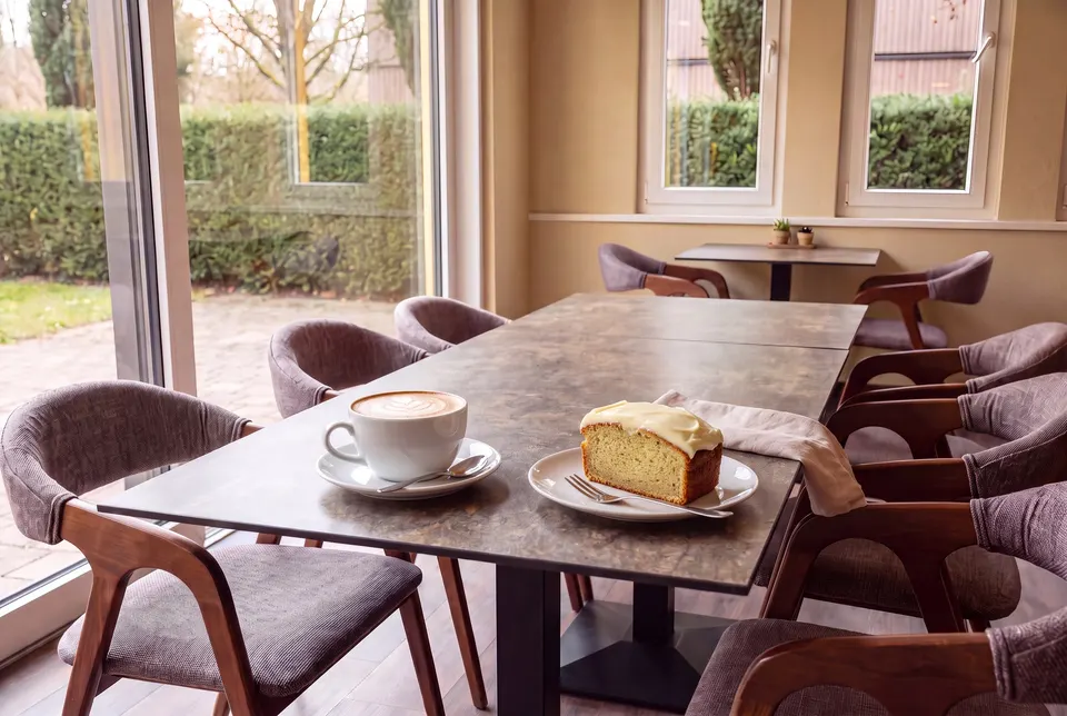 Cappuccino with latte art and a slice of banana cake with cream cheese frosting at the long table by the garden window (illustrative image)