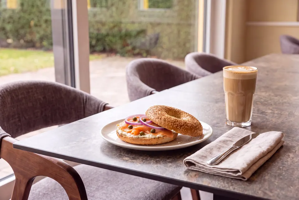 Salmon bagel with cream cheese, red onions and capers on a plate, with a latte macchiato beside it, at the café's window table (illustrative image)