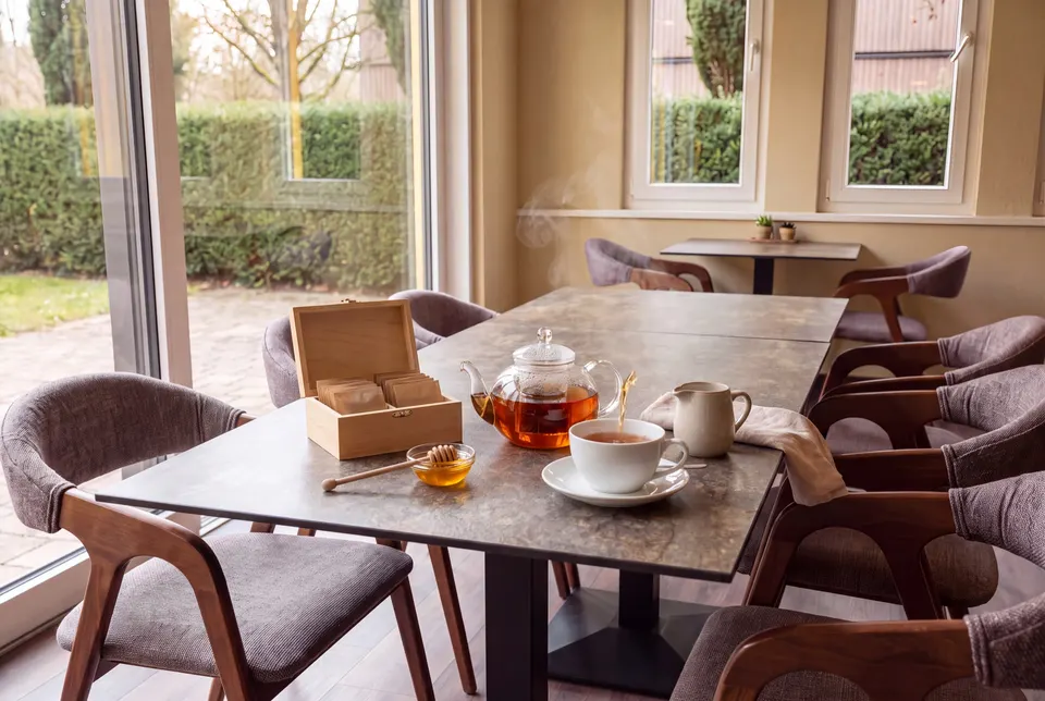 Glass teapot with black tea, a cup on a saucer and a small wooden box of tea bags on the table by the café's window (illustrative image)