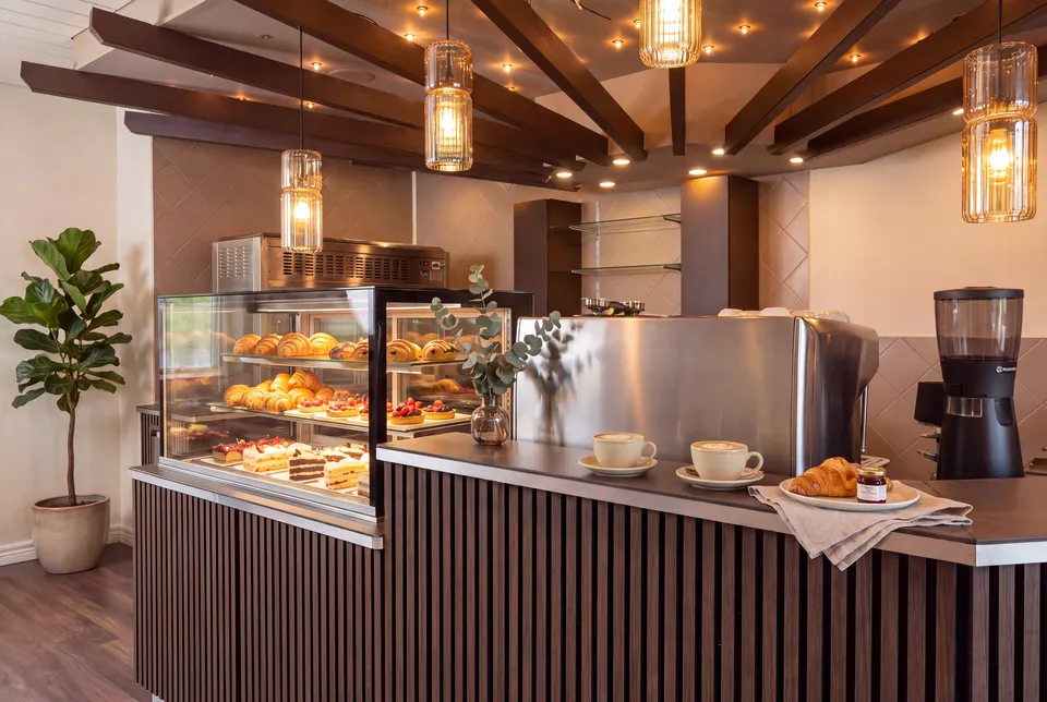 The café counter with a display of croissants, tarts and cakes, two cappuccinos and the espresso machine under wooden beams (illustrative image based on a photograph of the premises)