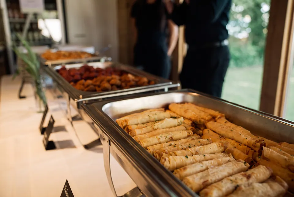 Catering buffet with chafing dishes full of golden cheese cigars, two people in black in front of a glass door in the background (illustrative image)