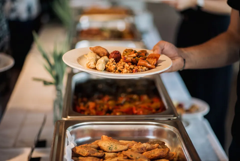 Guest at the buffet holding a plate with filled pastries, falafel and bulgur over a row of chafing dishes (illustrative image)