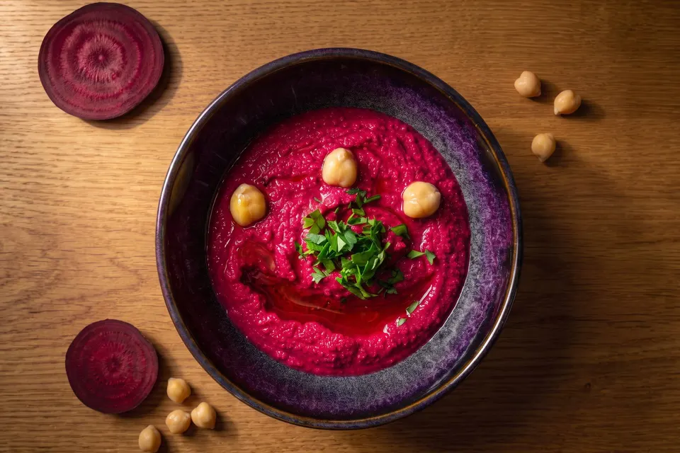 Pink beetroot hummus with chickpeas and parsley in a purple bowl, with slices of beetroot beside it (illustrative image)