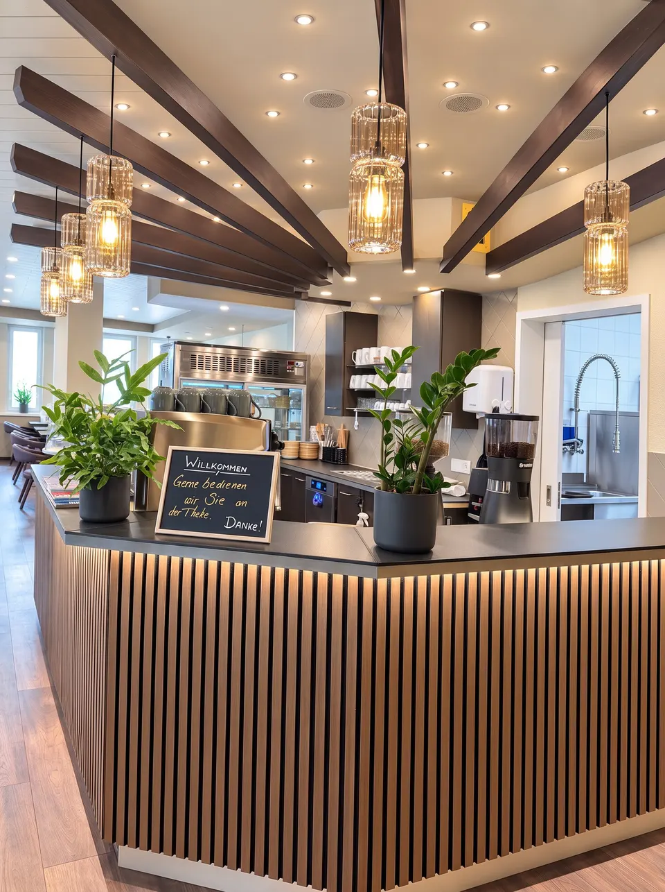 Counter with light wooden slats, plants and a welcome board, a coffee machine and the kitchen behind it, a fan of dark beams and pendant lights on the ceiling (illustrative image based on a photograph of the premises)