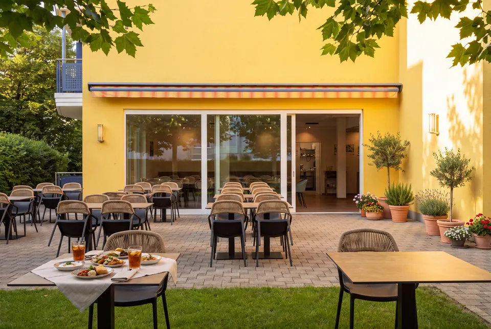 Terrasse vor der gelben Fassade mit Markise, Glasschiebetür, Olivenbäumchen, Lavendel und gedecktem Tisch auf dem Rasen (Symbolbild auf Basis einer Aufnahme des Hauses)